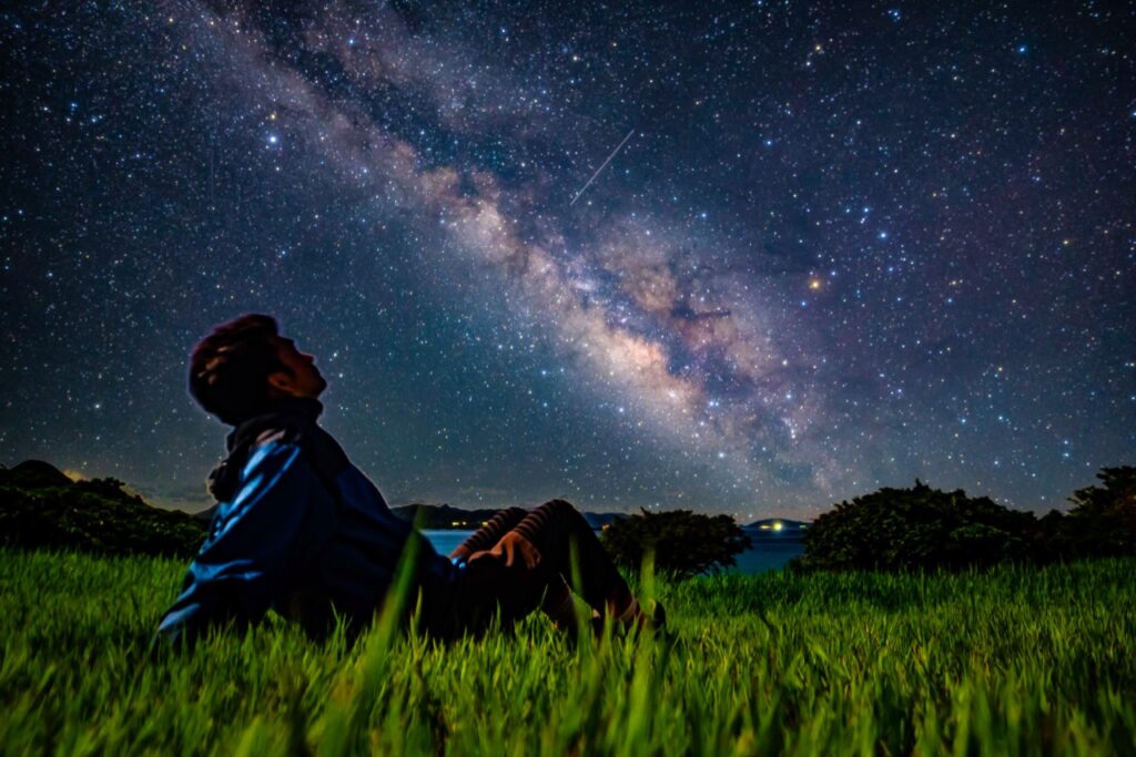 Man grassfield sitting milky way looking up at Nagareboshi no Oka stargazing spot, Ishigaki Island